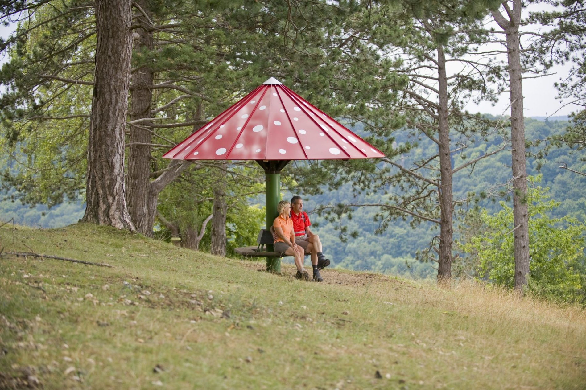 Zwei Wanderer sitzen an einer Sitzgelegenheit mit einem roten, pilzförmigen Dach.