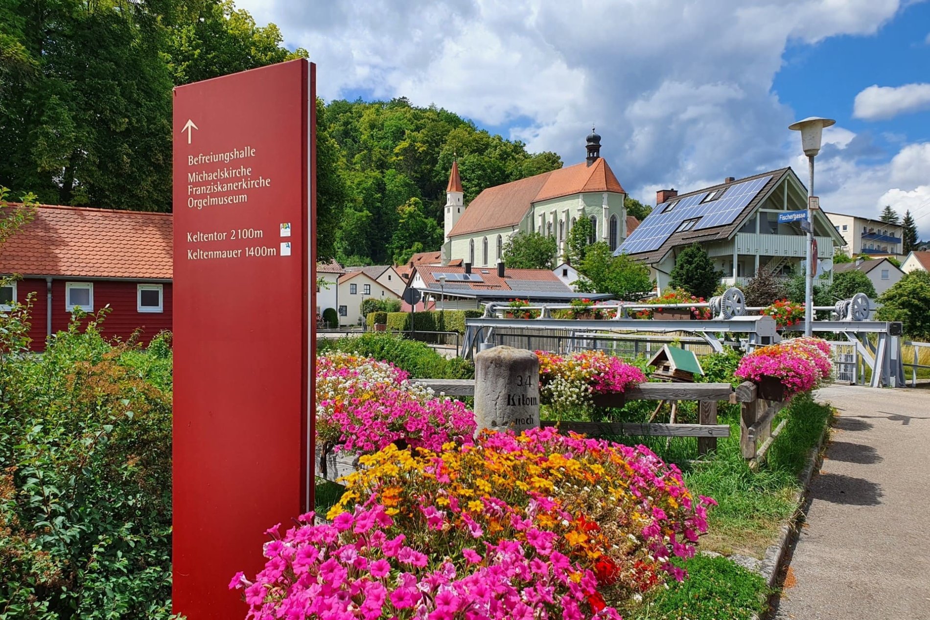 Links ist ein Hinweisschild vor einem bunten Blumenmeer, das auf das Orgelmuseum Kelheim hinweist. Die Kirche , in dem sich das Orgelmuseum befindet ist im Hintergrund sichtbar.