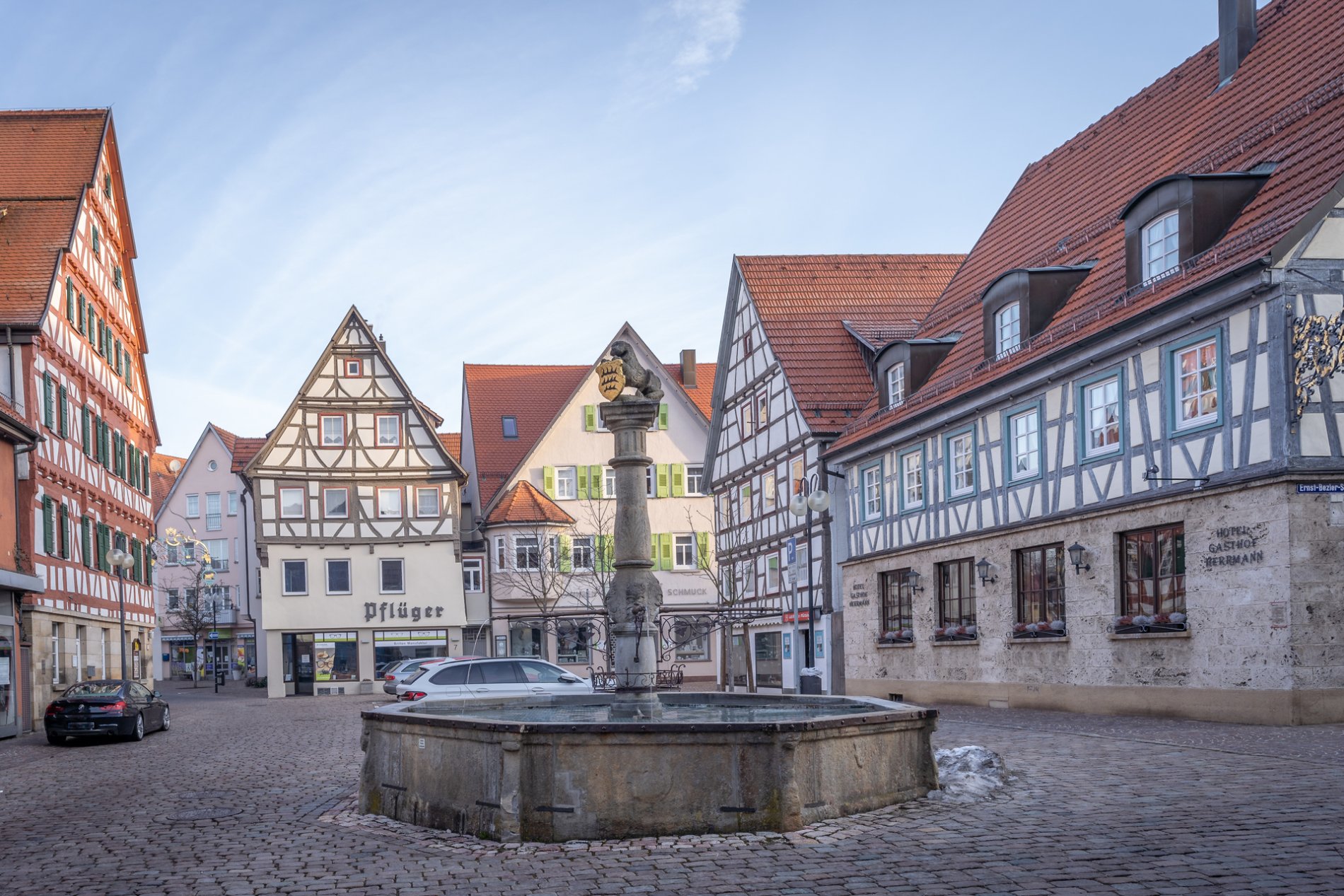 Der Marktplatz mit Marktbrunnen in Münsingen im Biosphärengebiet Schwäbische Alb. Ein großer Platz mit einem Brunnen in der Mitte an dessen Spitze ein Tier aus Stein und ein Wappen ist. Ringsherum sind Fachwerkhäuser.