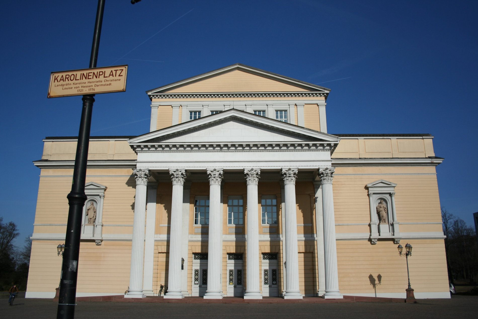 Classicist style building, made of yellowish stones with a white proticus in front of the main building with 6 white columns.