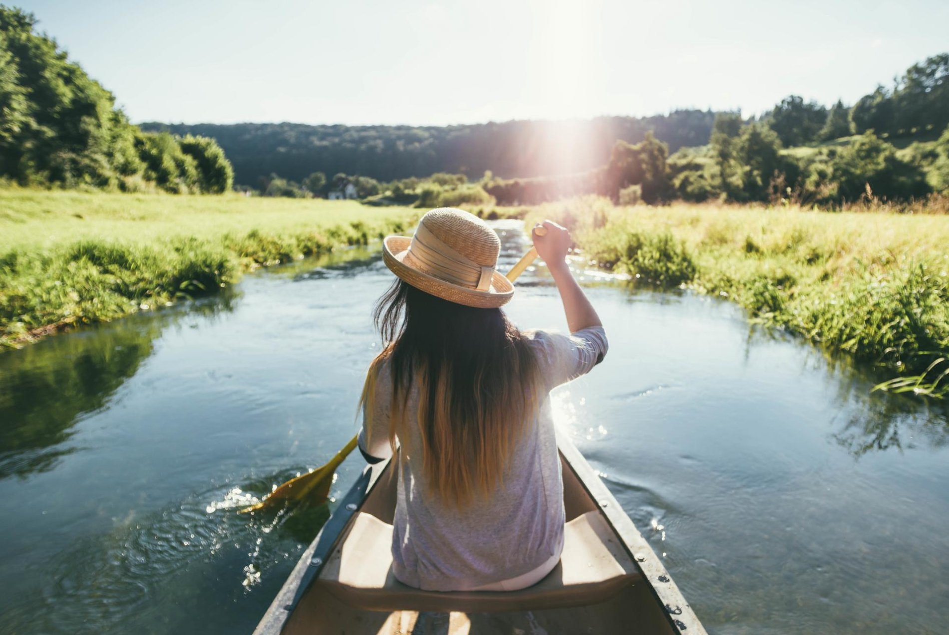 Eine Person sitzt vorne in einem Kanu und paddelt ruhig durch das Wasser. Sie trägt einen Sonnenhut. Links und rechts ist das dicht bewucherte Ufer. Die Sonne strahlt.