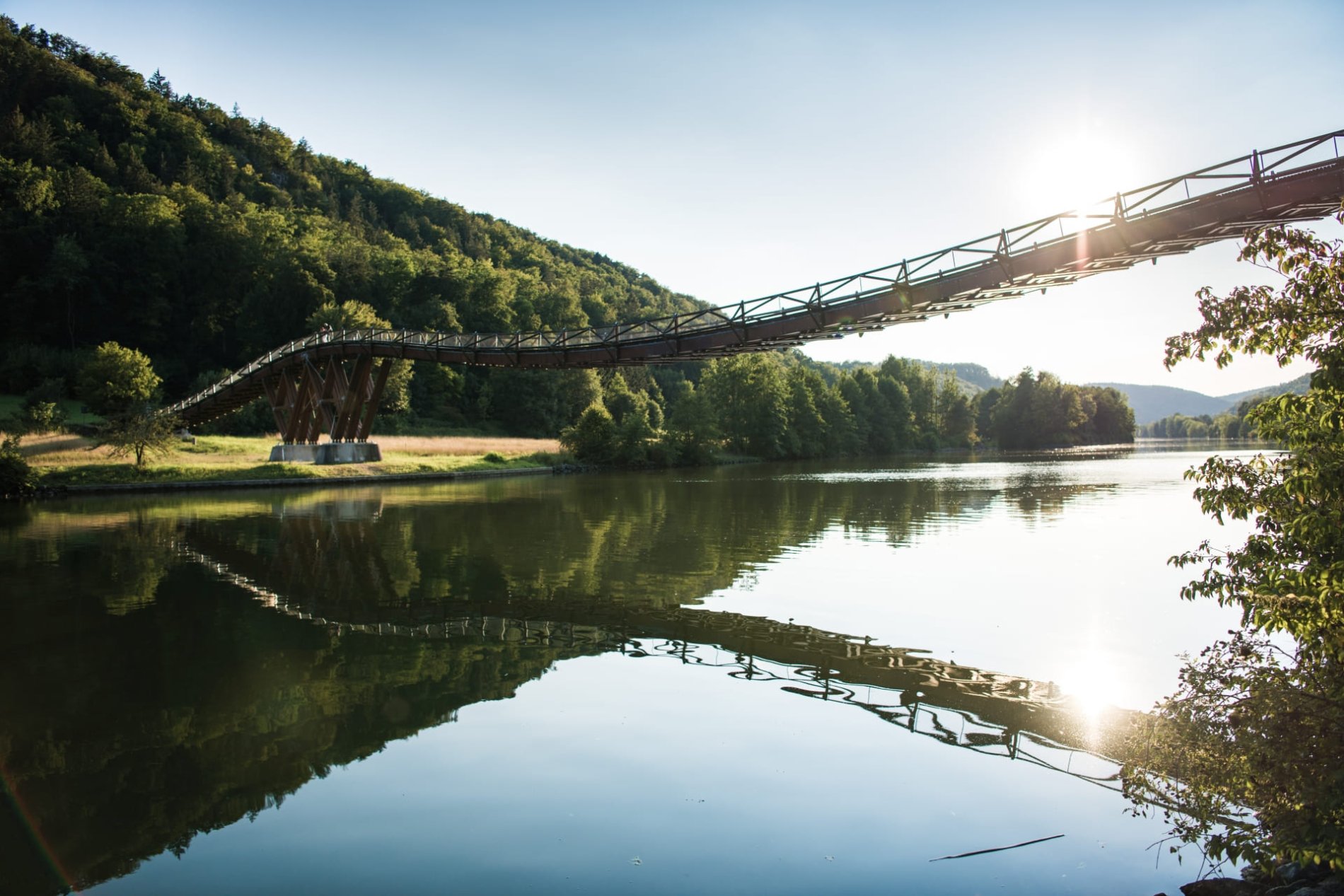 Im Wasser des Main-Donau-Kanals spiegelt sich die Holzbrücke Tatzlwurm. Auf der linken Seite befindet sich der Wald. Tief steht die Sonne im Hintergrund.