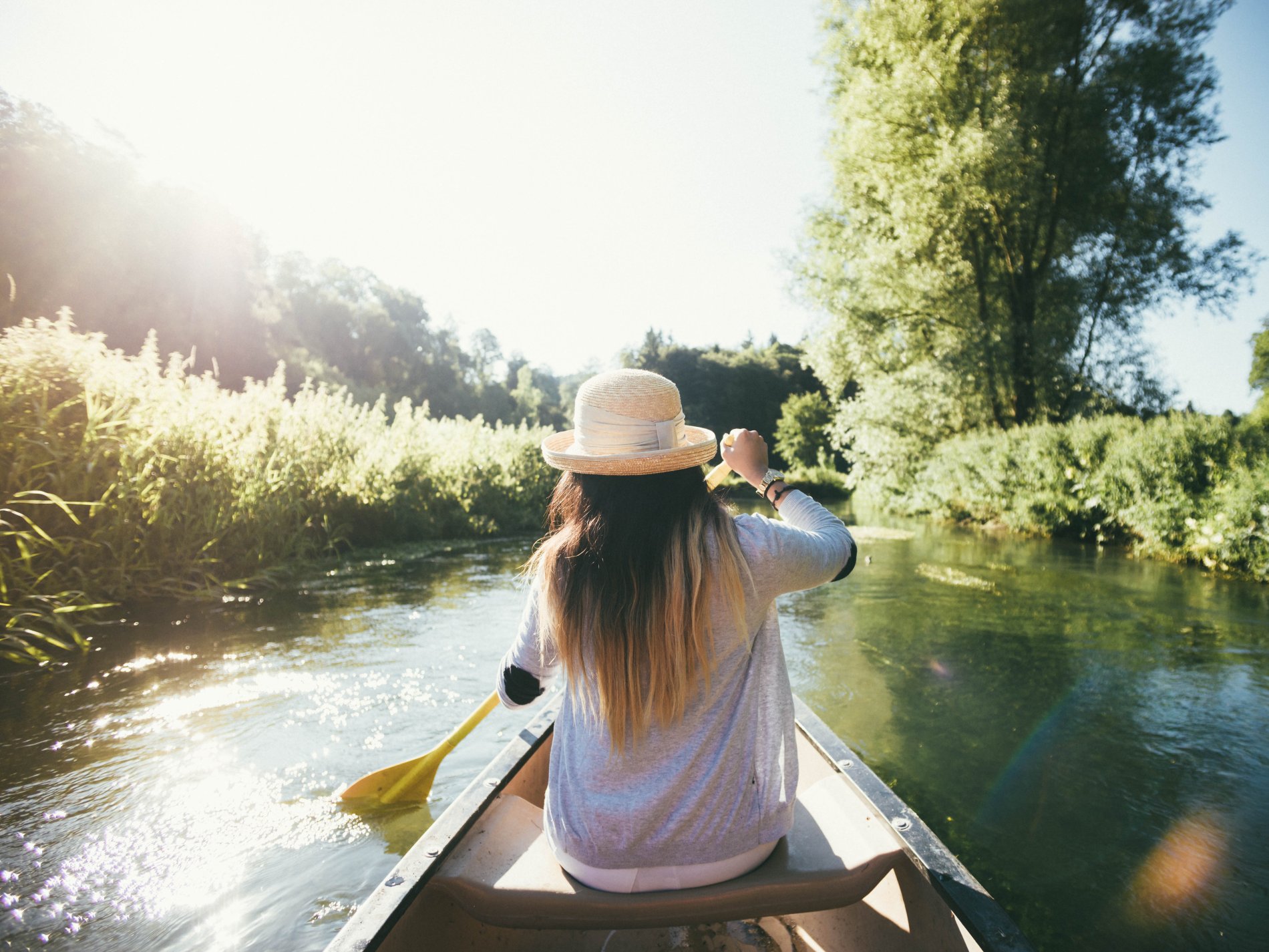 Eine Frau sitzt in einem Kanu und paddelt entlang einem Fluss. Vom Ufer ragen Pflanzen über den Fluss. Sie trägt einen Sonnenhut.