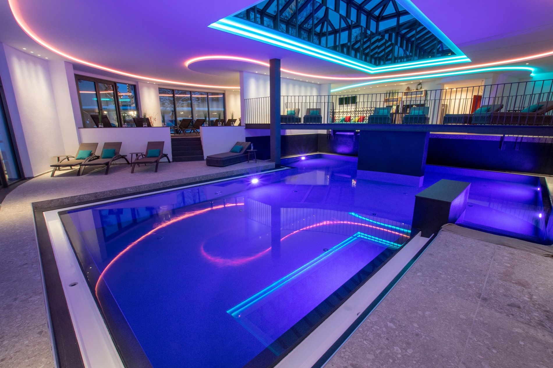 Ein stimmungsvoll in blau beleuchteter Indoor-Pool- Links stehen einige Liegen. Dahinter ist eine große Terrasse mit weiteren Liegen. An der Decke sind bunte LEDs und ein Panorama-Dach mit großen Fenstern.