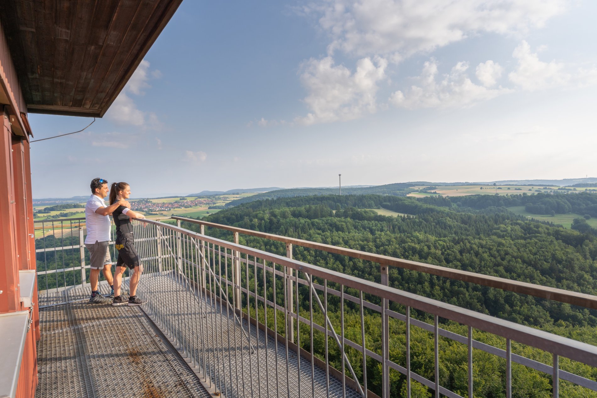 Zwei Personen stehen auf einer Aussichtsplattform aus Metall und lehnen sich an das Metallgeländer. Ringsherum ist ein weiter Ausblick über Wälder, Wiesen und ein Dorf am Horizont. Daneben ist eine rote Wand des Turms.