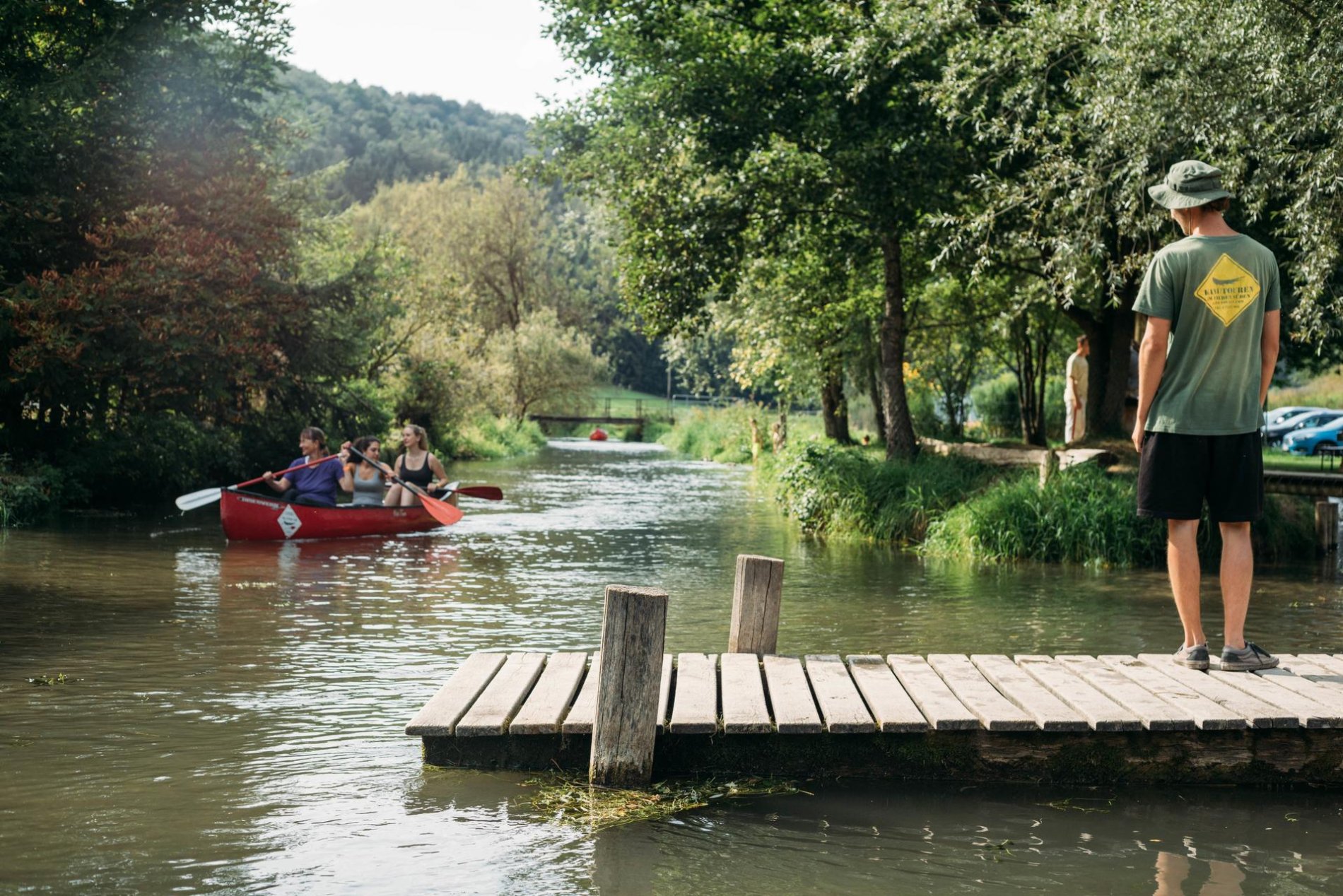 Auf einem Steg an einem Fluss steht eine Person mit Hut und blickt zu einem Kanu mit drei Personen, die gerade vorbei paddeln. Am Ufer sind Bäume und Pflanzen.