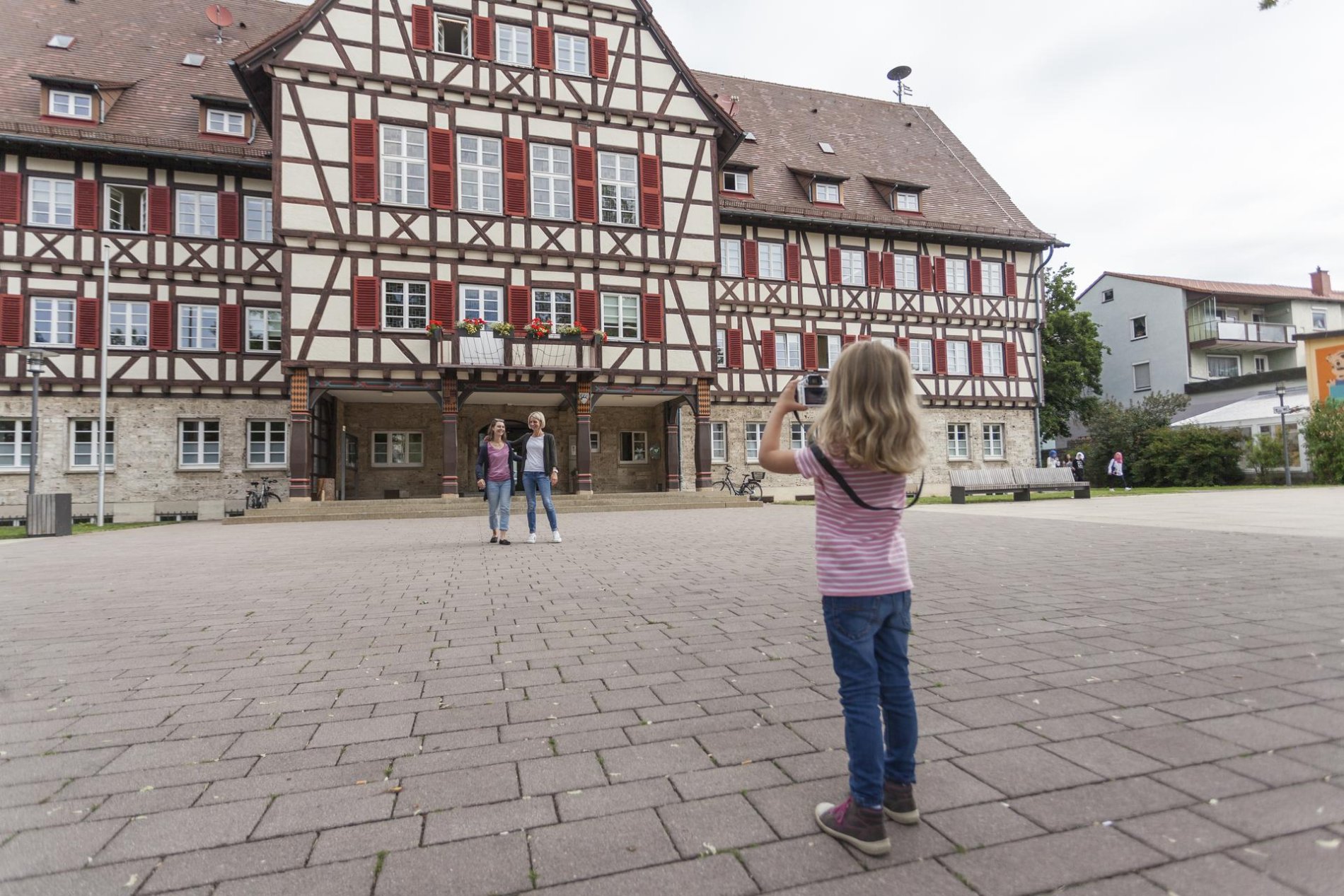 Das Rathaus in Münsingen im Biosphärengebiet Schwäbische Alb. Ein Kind steht mit einer Digitalkamera auf einem Platz und richtet diese in Richtung von zwei Erwachsenen, die sich vor einem historischen Fachwerkhaus aufstellen.