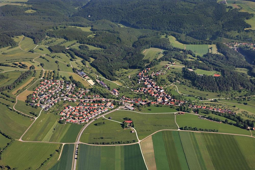 Ein Luftbild eines Dorfs, das von einem weiten Straßennetz durchzogen ist. Ringsherum sind Felder, Wiesen und Wälder. Am rechten Bildrand ist noch ein weiteres Dorf.