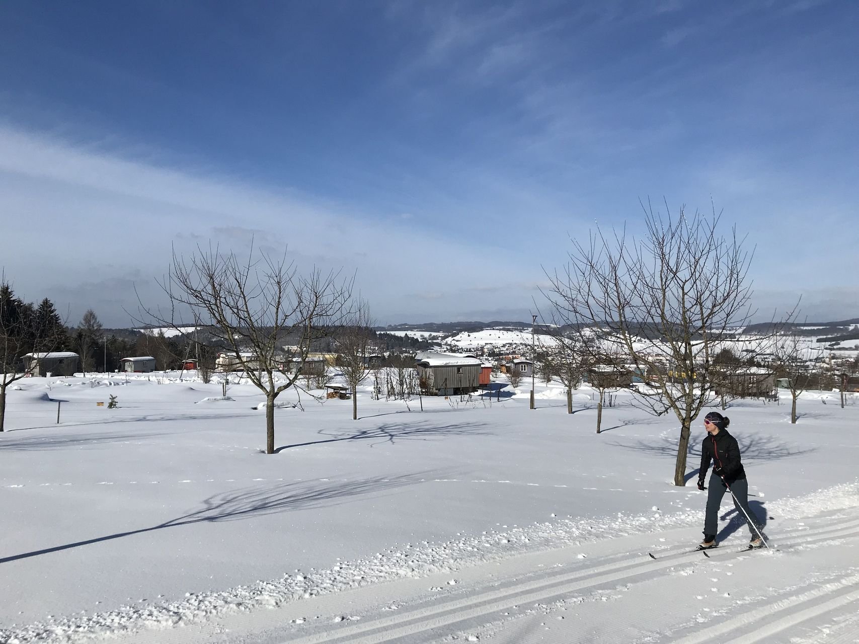 Eine Person läuft mit klassischen Langlaufski in Spurrillen durch eine verschneite Landschaft. Im Hintergrund sind einige Schäferwagen und Bäume. Die Sonne scheint.