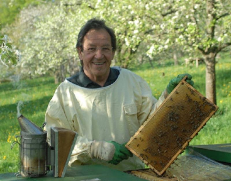 Imkerei und Honigverkauf Gekeler in Münsingen im Biosphärengebiet Schwäbische Alb. Ein*e Imker*in hält eine Bienenwabe in den Händen und lächelt in die Kamera. An der Bienenwabe hängen mehrere Bienen. Im Hintergrund sind blühende Bäume.
