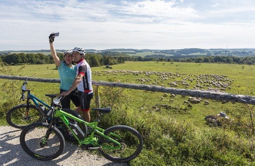 Zwei Radfahrer*innen stehen mit dem Rücken zu einer Schafherde und machen mit dem Handy ein Selfie. Der Ausblick reicht über die weite Albhochfläche.