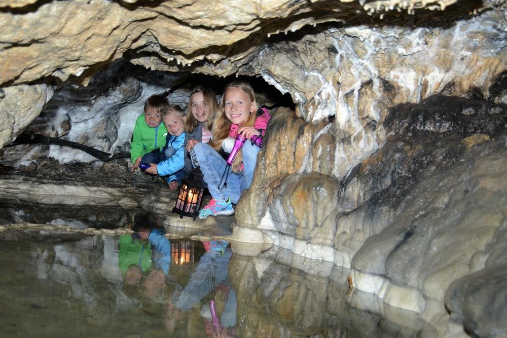 Vier Kinder sitzen in der Hocke in einer Nische in einer Höhle. Zwei haben eine Taschenlampe, ein Kind hält eine Laterne. Vor ihnen ist Wasser und an der Decke bilden sich Tropfsteine.