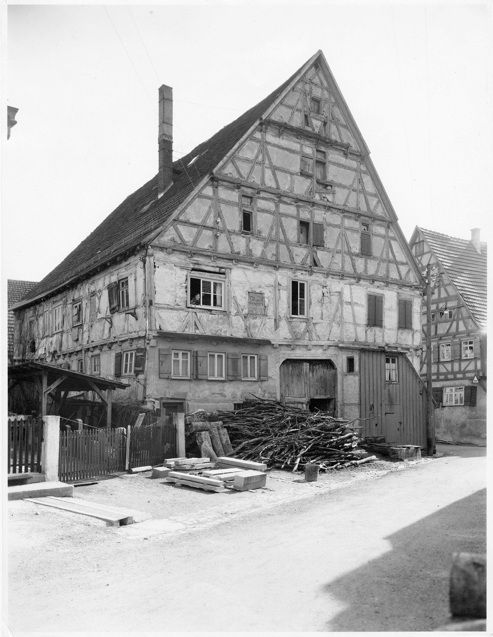Das Große Haus früher in Münsinge im Biosphärengebiet Schwäbische Alb. Ein Fachwerkhaus in schwarz weiß, dessen Fassade schon leicht zerfallen ist. Links davon ein Gartenzaun, davor ein Holzstapel.