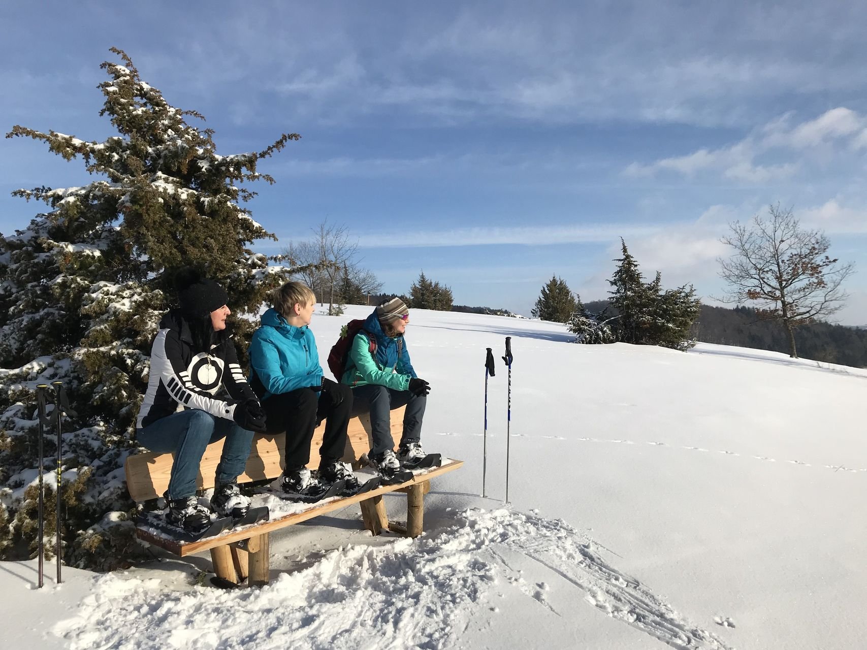 Drei Schneeschuhwanderer*innen sitzen auf einer Holzbank und schauen mit einem Lächeln in die Ferne. Ihre Wanderstöcke stecken im Schnee. Es ist sonniges Wetter und etwas bewölkt. Im Hintergrund sind vereinzelt Bäume.