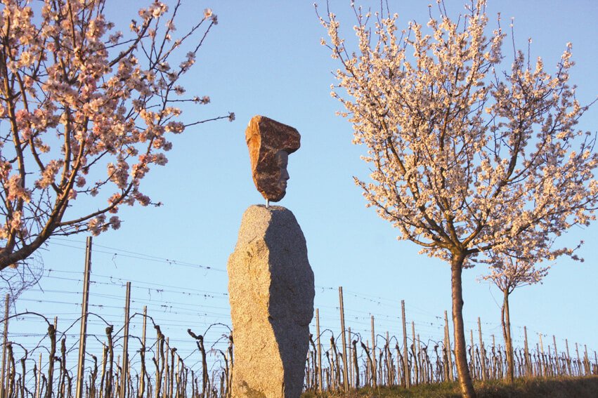 Eine Steinskulpur aus zwei Felsbrocken, stellen einen Weingott dar, im Hintegrund sind Weinreben zu sehen, links und rechts stehen blühende Mandelbäume.