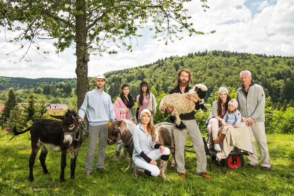 Mehrere Personen auf einer grünen Wiese mit einem Esel, Ponny und Schaf. Sie posieren für die Kamera. Es ist sonnig. Hinter ihnen steht ein Baum.