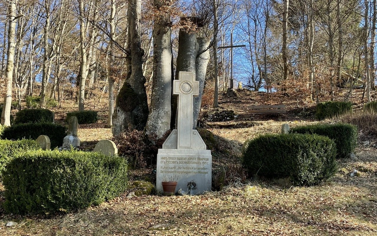 Der Friedhof Gänsewag auf dem ehemaligen Truppenübungsplatz in Münsingen im Biosphärengebiet Schwäbische Alb. Ein steinernes Kreuz mit einer Innschrift daneben sind Buchshecken, im Hintergrund ein dicker an der Spitze abgesägter Baum und ein großes Kreuz