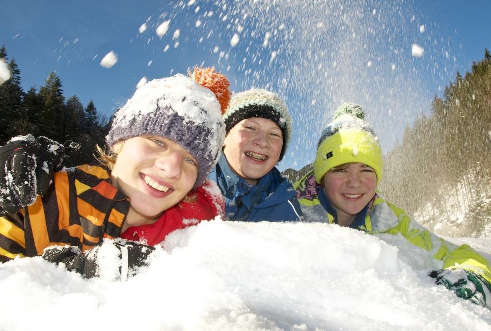 Drei Kinder mit bunten Winterjacken und Mützen lächeln in die Kamera während sie sich auf einem Schneehaufen abstützen. Von oben kommt noch mehr Schnee, der auf sie herabfällt. Im HIntergrund ist Wald. Der Himmel ist blau.