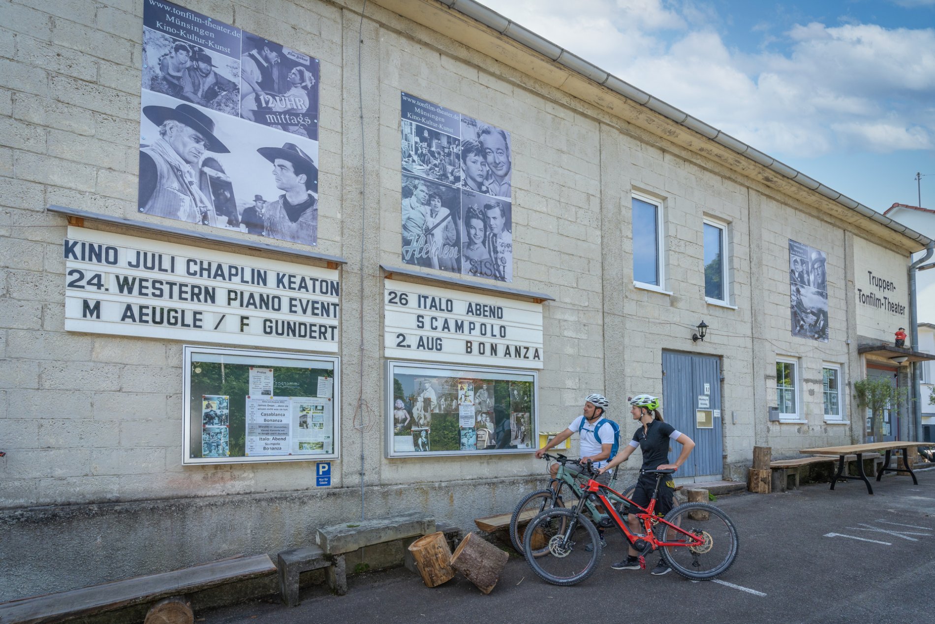 Zwei Radfahrer*innen stehen vor einem grauen Gebäude mit der Aufschrift Truppen- und Tonfilm-Theater. An der Wand sind mehrere Plakate in schwarz-weiß mit Filmklassikern. Sie betrachten die Tafel und den Schaukasten mit dem Programm.