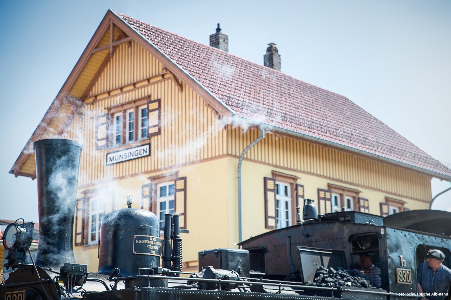 A steam locomotive stands in the foreground at Münsingen station. It is surrounded by steam clouds. In the background is the historic station building of Münsingen.