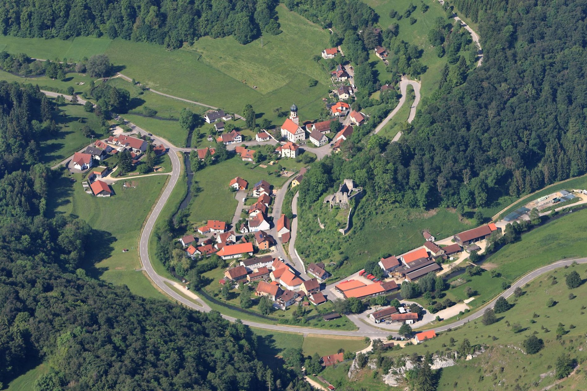 Ein Luftbild von einem Dorf, durch das ein Fluss fließt. Auf einem Hügel ist eine Burgruine.