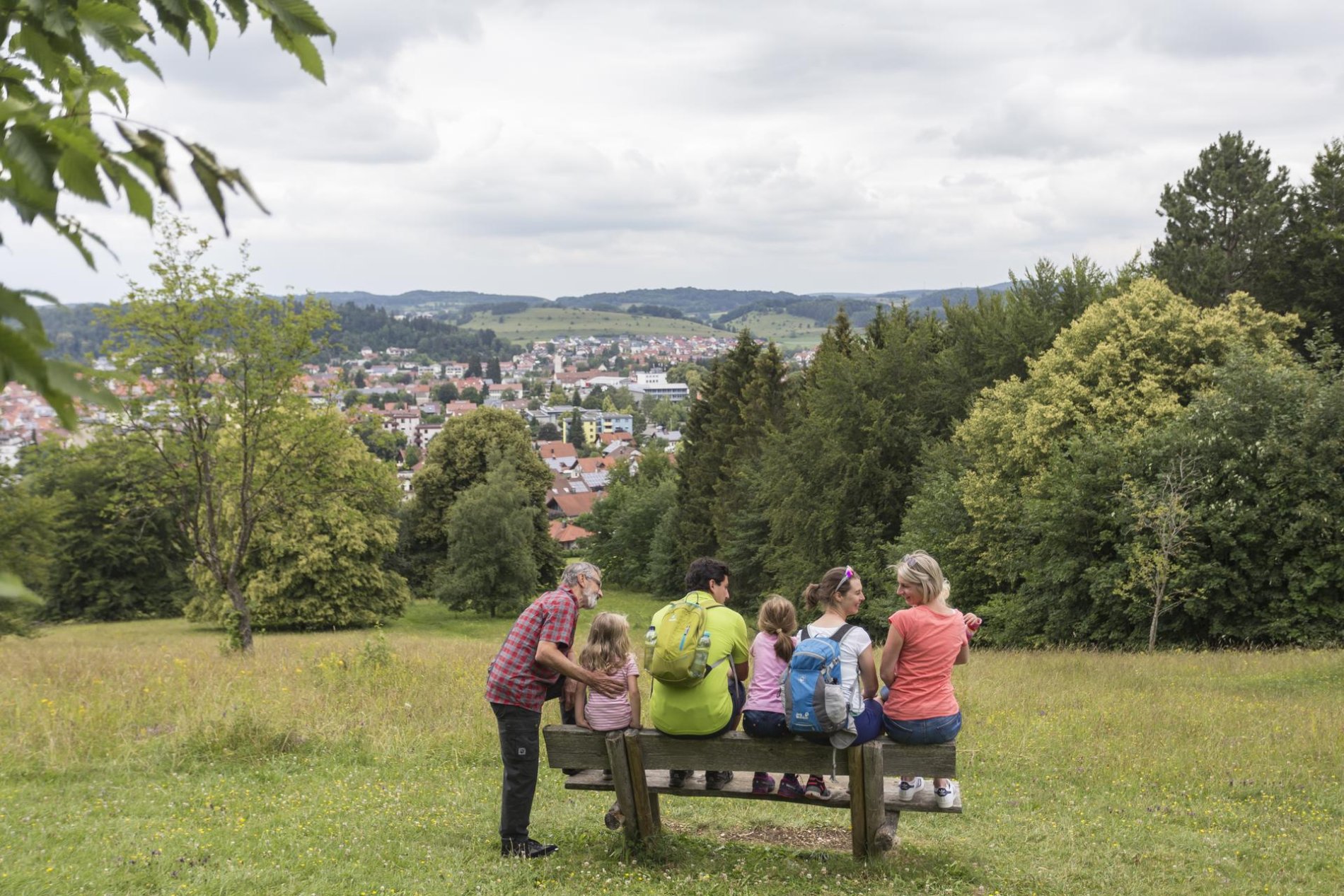Gruppenreise ins Biosphärengebiet Schwäbische Alb. Ein Tag in der Münsinger Innenstad. Auf einer Holzbank sitzen mehrere Personen und Kinder. Vor ihnen ein weiter Ausblick über eine Stadt. Ringsherum ist Natur. Der Himmel ist bewölkt.t