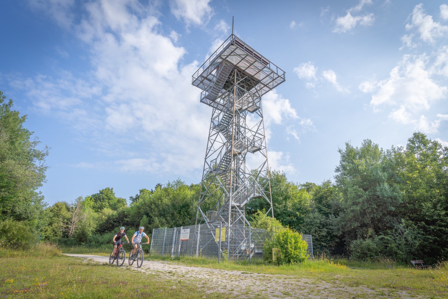 Turm Waldgreut auf dem ehemaligen Truppenübungsplatz in Münsingen im Biosphärengebiet Schwäbische Alb. Ein hoher Stahlgitterturm, der von einem Metallzaun umgeben ist. Davor ist ein Schotterweg auf dem zwei Radfahrer*innen fahren.