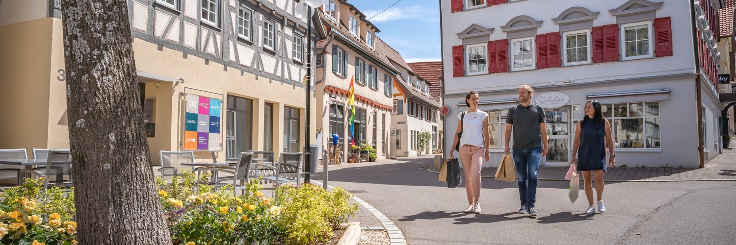 Drei Personen laufen lächelnd mit Tüten und Taschen durch eine Innenstadt, die von Fachwerkhäusern und kleinen Läden geprägt ist. Es ist schönes Wetter und warm.