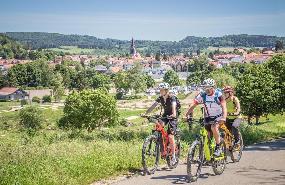Drei Radfahrer*innen fahren mit E-Bikes in kurzen Radklamotten versetzt auf einem Teerweg. Im Hintergrund ist eine Stadt, ein Bikepark und vereinzelt Wohnmobile.