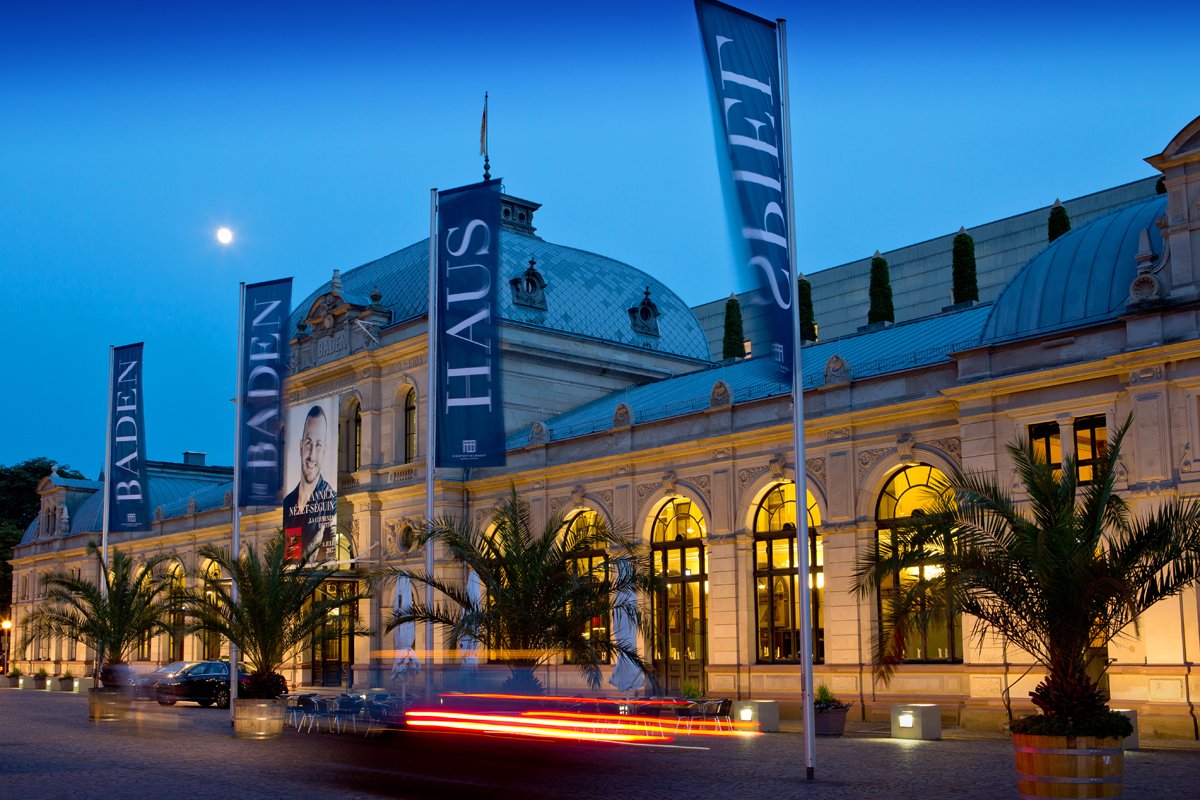 Gebäude des Festspielhaus Baden-Baden beleuchtet bei Nacht mit blauen wehenden Fahnen. Das Banner in der Mitte zeigt den Dirigenten Yannick Nézet-Séguin.