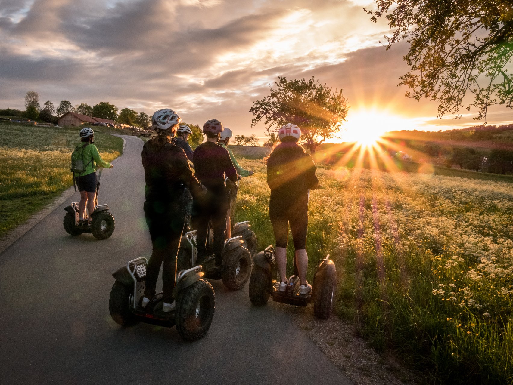Segway Touren rund um Münsingen im Biosphärengebiet Schwäbische Alb. Eine Grupee steht mit Segways auf einem Teerweg und beobachten den Sonnenuntergang. Die Sonne wirft Strahlen über blühende Sommerwiesen. Alle tragen Helme.