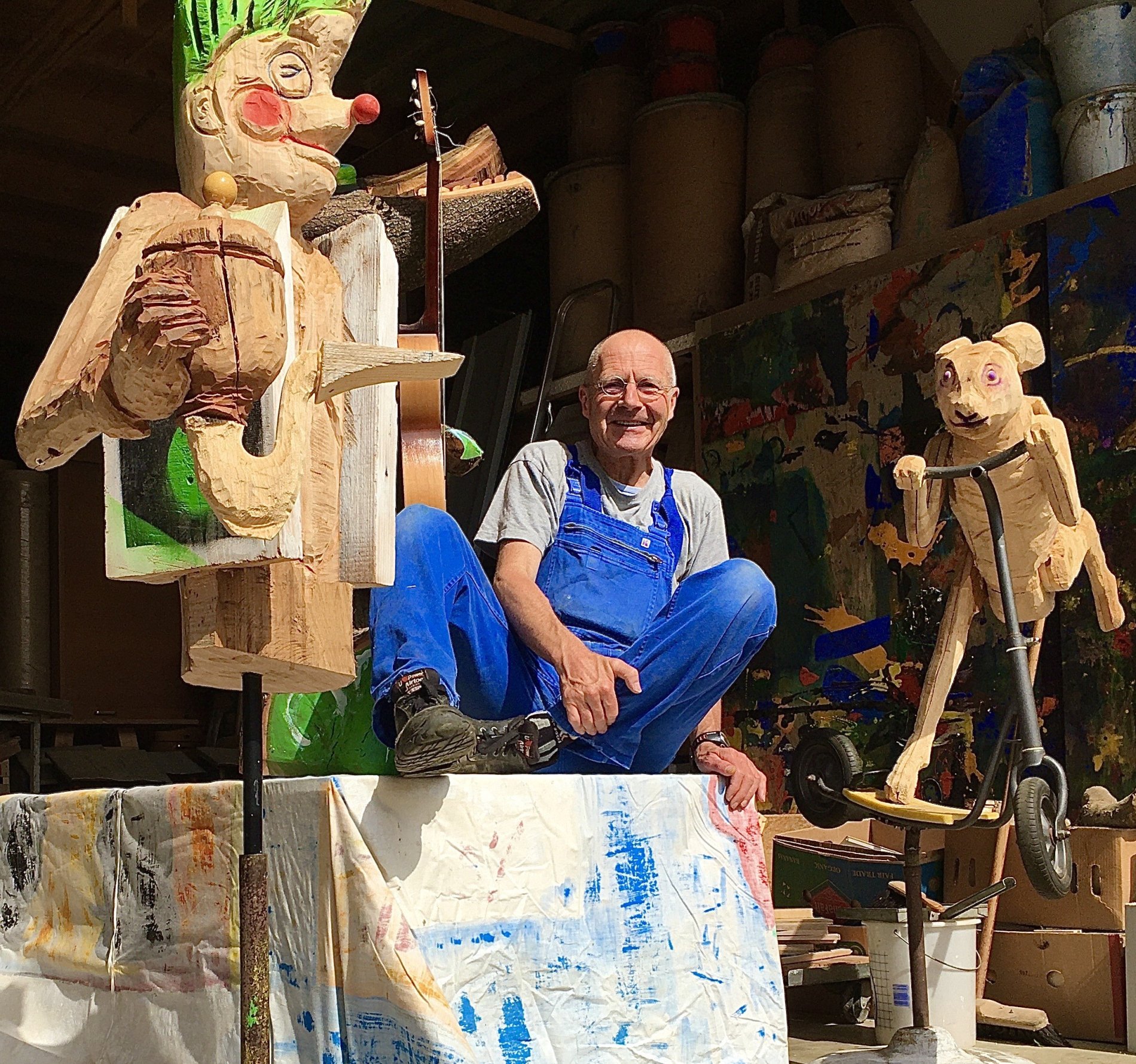 Mann sitzt neben Holzkunstwerken mit blauer Latzhose