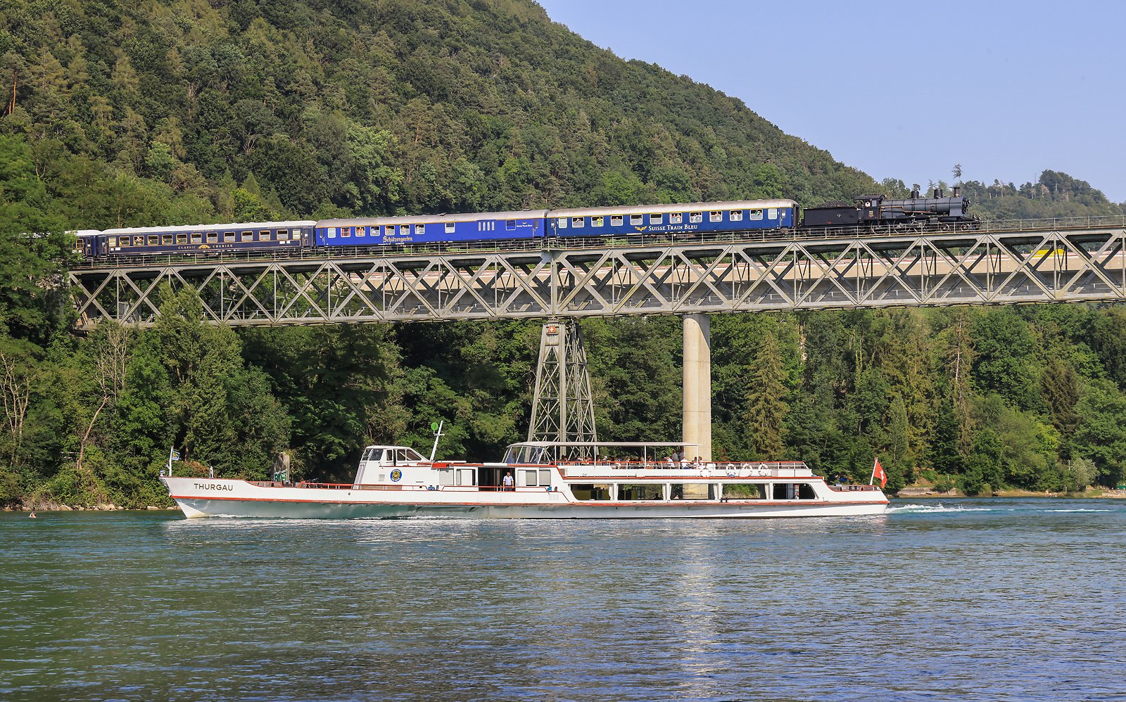Dampflok fährt über eine Rheinbrücke, unter der ein weißes Passagierschiff fährt.