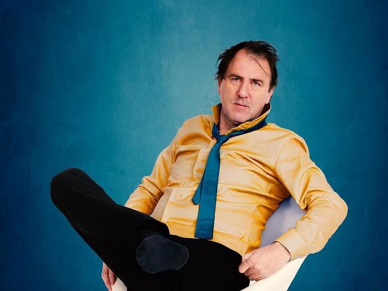 Kabarettist Stefan Waghubinger vor blauem Hintergrund in einem Sessel