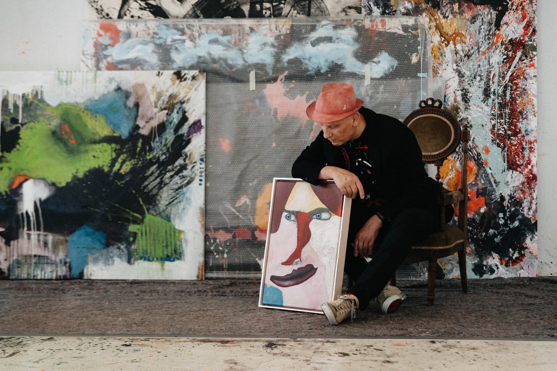 Dietmar Schönherr sitzt vor einer Wand mit Gemälden und begutachtet eines seiner Werke