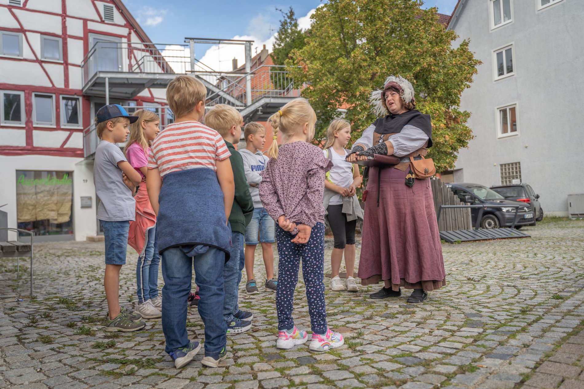 Kinderführung mit dem Marktweib Marga in Münsingen im Biosphärengebiet Schwäbische Alb. Eine Person in einem mittelalterlichen Kostüm steht neben mehreren Kindern und erzählt etwas. Im Hintergrund sind Häuser und Bäume.