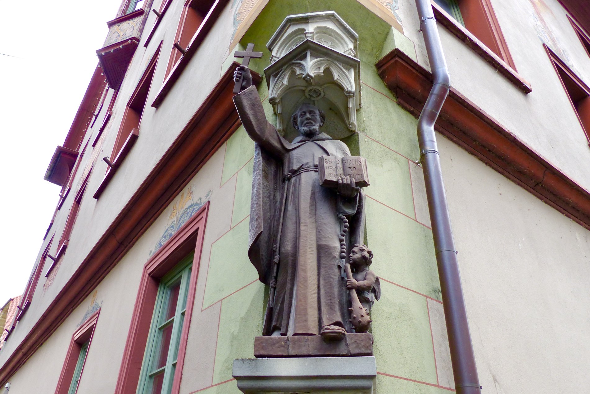 Holzstatue des Heiligen Fidelis an der Nordostecke des Fidelishauses. In der erhobenen rechten Hand hält der Heilige ein Kreuz. Zu seinen Füßen steht ein kleiner Engel mit mit dem Attribut des Heiligen Fidelis, einer Keule.