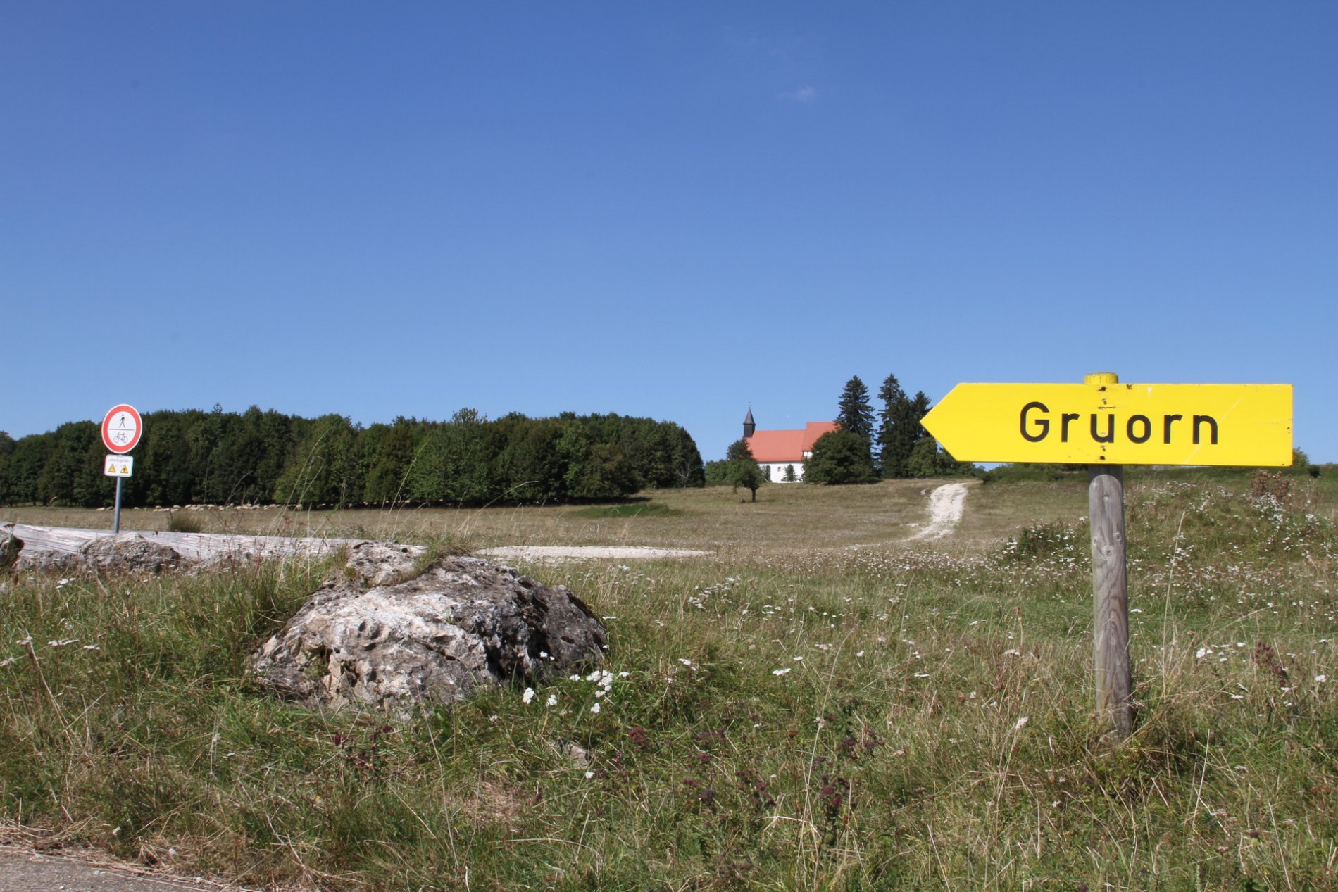Im Vordergrund ist ein gelbes Holzschild mit der Aufschrift Gruorn. Daneben ist ein Fels. Im Hintergrund ist ein kleiner Hügel und eine Kirche zu der ein kleiner Schotterweg führt. Der Himmel ist blau.