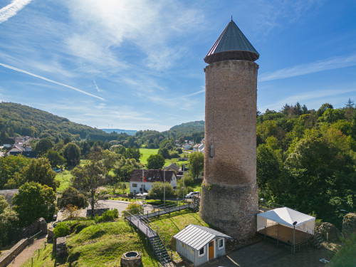 Das Bild zeigt den gesamten Bergfried der Burg Nohfelden und einen Blick in die hügelige Mittelgebirgslandschaft. Es ist ein sonniger Tag, Bäume und Wälder im Hintergrund sind begrünt. Am Fuß des Burgturms sind ein Zelt und eine Hütte für Veranstaltungen