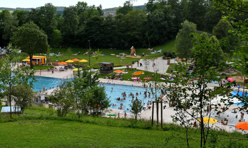 Ein Freibad mit einem großen Becken, einer Wasserrutsche, einem kleinen Kinderbecken, einem Wasserspielplatz, vielen Liegewiesen, einem Kiosk und weiteren Spielmöglichkeiten. Überall tummeln sich Leute. Im Hintergrund sind Bäume.