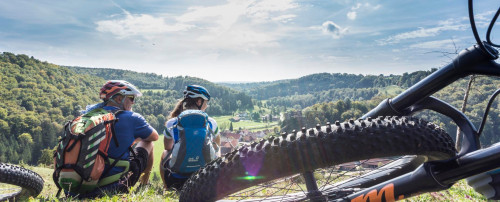 Münsinger e-Bike  Tour 1: eine Radtour von Münsingen durch das Biosphärengebiet Schwäbische Alb. Zwei Radfahrer*innen sitzen auf einem Hügel und blicken lächelnd ins Tal. Im Vordergrund liegt ein Mountainbike.