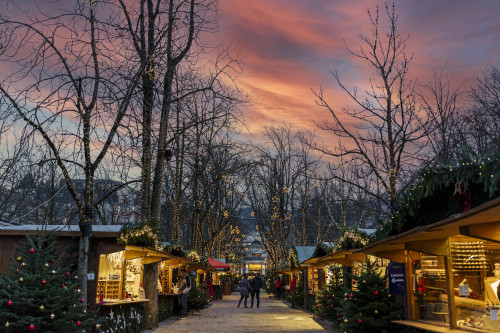 Ein Pärchen schlendert am Baden-Badener Christkindelsmarkt entlang, vorbei an traditionellen Holzbuden, während der Himmel in beeindruckenden Rosé- und Rottönen durch den Sonnenuntergang erstrahlt.