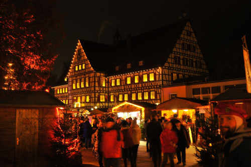 Der Weihnachtsmarkt in Münsingen im Biosphärengebiet Schwäbische Alb. Ein festlich beleuchteter Platz mit einem historischen Fachwerkgebäude im Hintergrund. Auf dem Platz stehen viele Hütten und mehrere Personen.