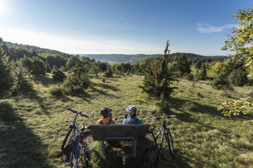 Die e-Bike Tour 5 führt von Münsingen nach Schelklingen durch das Biosphärengebiet Schwäbische Alb. Zwei Radfahrer*innen sitzen auf einer Holzbank und genießen den Ausblick über die weite Alblandschaft mit viel Wald. Neben ihnen stehen zwei Fahrräder.