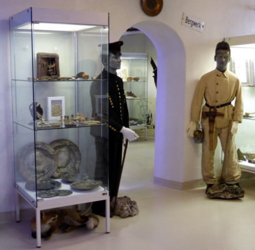 Sichtbar ist ein Teil des Museums, mit einer Vitrine mit Ausstellungsstücken und nebendran zwei Puppen in typischen Bergwerks-Kleidern.