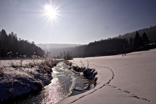 Ein Fluss, der teilweise gefroren ist, zieht sich durch eine Schneelandschaft. Die Sonne scheint und im Hintergrund ist Wald.