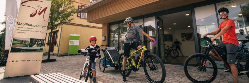 Zwei Erwachsene und ein Kind stehe in kurzer Radkleidung mit ihren Fahrrädern vor einem Gebäude. Die beiden Erwachsenen haben E-Bikes und einer einen Kinderfahrradanhänger. Vor dem Gebäude steht ein Schild mit der Aufschrift Mobilitätszentrum.