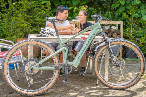Die Radtour Berg Bier-Tour von Münsingen nach Ehingen zur Berg Brauerei im Biosphärengebiet Schwäbische Alb. Zwei Personen sitzen an einem Tisch im Garten und stoßen mit zwei gefüllten Gläser an. Sie grinsen. Vor ihnen lehnt ein e-Bike am Tisch.
